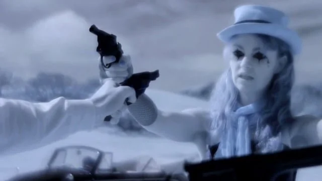 A woman with pale skin and long, wavy hair, wearing a light-colored hat and scarf, pointing a gun at the camera inside a vehicle in a snowy landscape.