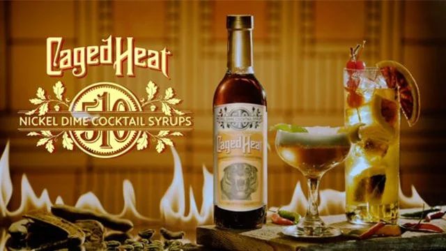 A bottle of Caged Heat nickel dimes cocktail syrup, a cocktail glass, and a tall glass with a drink garnished with fruit slices and a cherry, set against a warm background with flames.