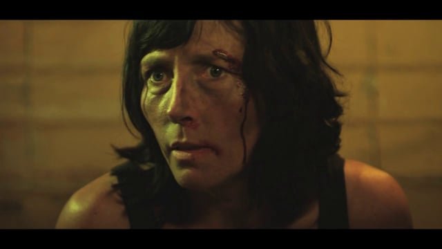 A woman with long dark hair, bruised face, and serious expression looking to the side in a dimly lit room.