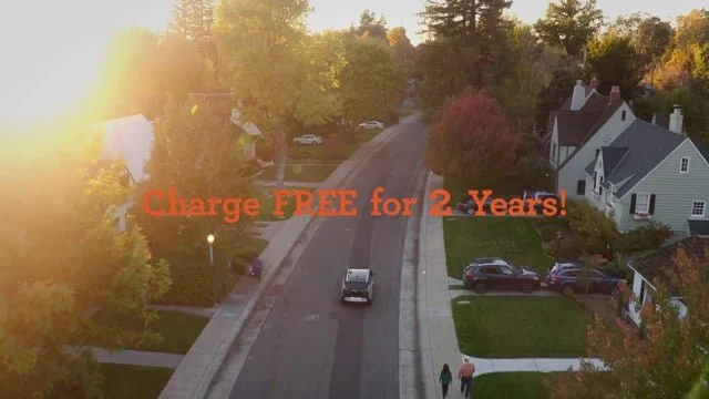 Aerial view of a suburban street during sunset with colorful trees, houses, cars parked on the street, and two people walking on the sidewalk. Overlaid text reads, 'Charge FREE for 2 Years!'