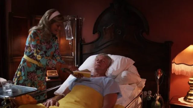 A woman in a colorful floral dress is leaning over an elderly man lying in bed, who is covered with a yellow blanket. The room has warm lighting and wooden walls, with a headboard and a lamp visible on the bedside table.