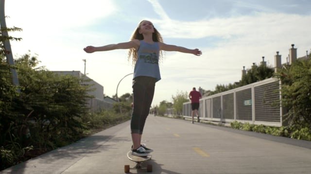 A girl with long hair skateboarding on a trail with her arms outstretched and a smile on her face, under a bright sky.