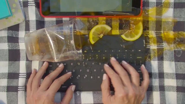 A person typing on a small black keyboard with a checkered tablecloth, a plastic cup, and lemon slices nearby.