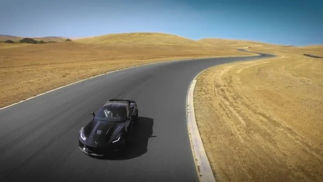 A black sports car driving on a curved mountain road surrounded by yellow grass and hills under a clear blue sky.