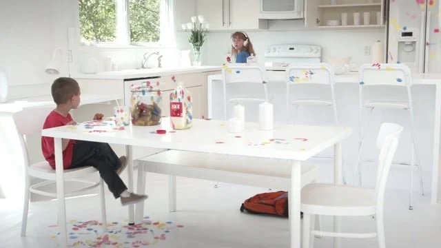 A bright, white kitchen with two children sitting at a dining table, and a girl playing in the background.