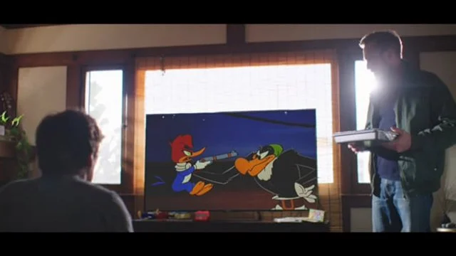 Two young men are in a room watching a cartoon, with one seated and the other standing holding a box of pizza. The cartoon on the TV features Donald Duck and a character dressed as a pirate.