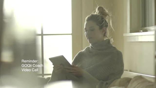 A woman with a bun hairstyle sitting on a bed, holding a tablet, in a sunlit room with a window in the background.