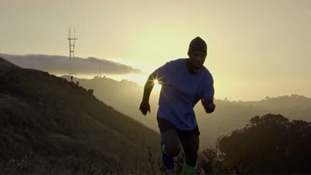 A man running uphill outdoors during sunset or sunrise, wearing a hat and athletic clothing, with hills and a transmission tower in the background.
