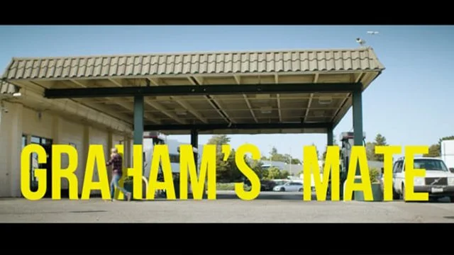 A large yellow text overlay that reads 'GRAHAM'S MATE' in front of a carport or covered parking area with cars parked underneath and a building on the left.