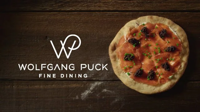 A pizza with pepperoni, black olives, and chopped herbs on a wooden table next to a Wolfgang Puck Fine Dining logo.
