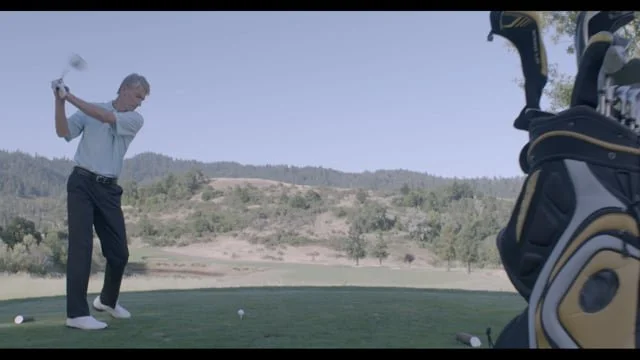 A person is swinging a golf club on a golf course, preparing to hit a ball. A golf bag filled with clubs is visible in the foreground on the right side of the image.