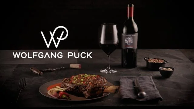 A dinner setting featuring a glass of red wine, a bottle of red wine with a Wolfgang Puck label, a cooked steak on a plate, a gray napkin with a fork and knife, and small bowls of seasoning or sauce on a dark surface.