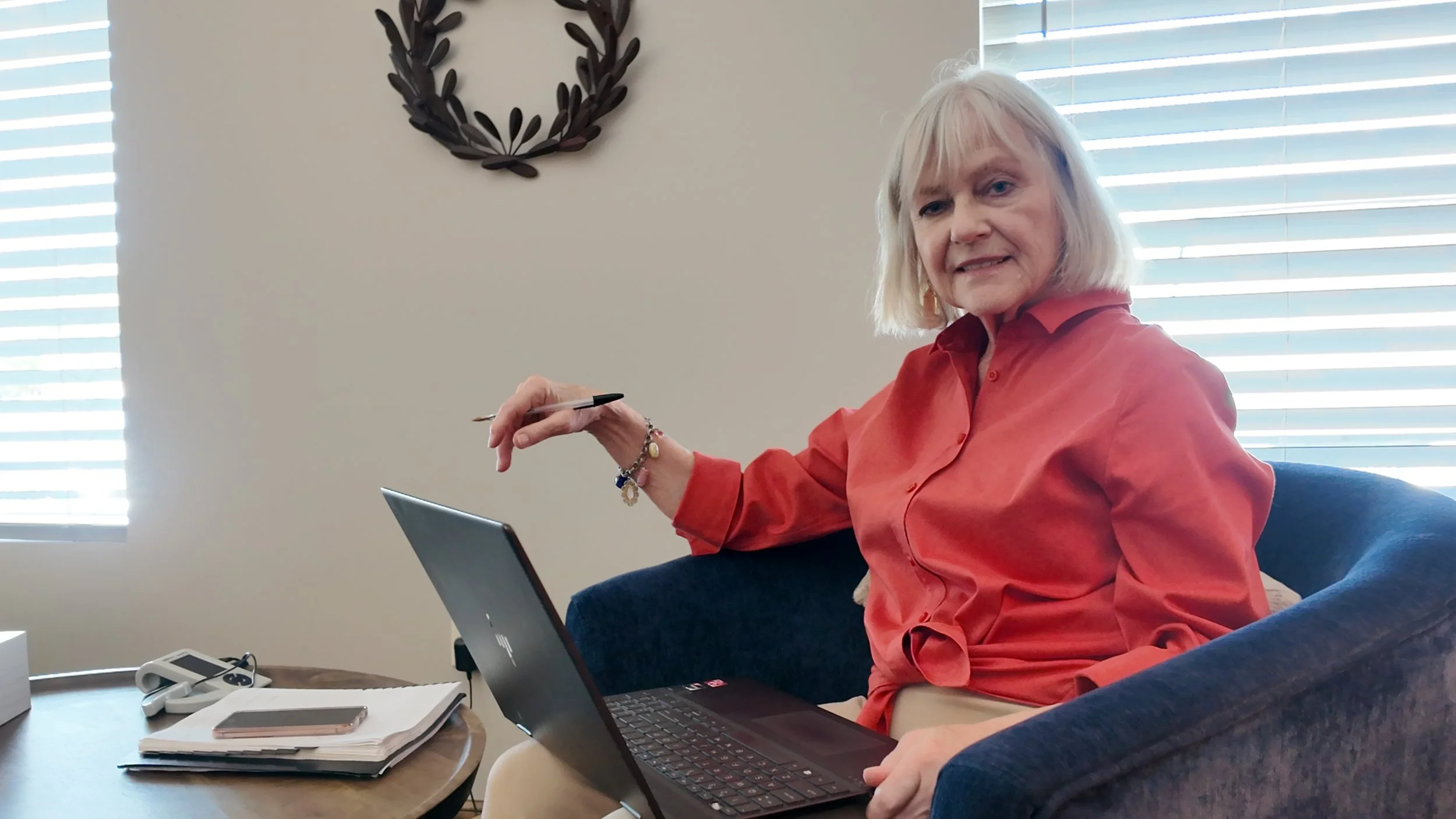A woman with blonde hair in a red blouse sitting at a desk with a laptop, pointing at the screen, with blinds on the window behind her.