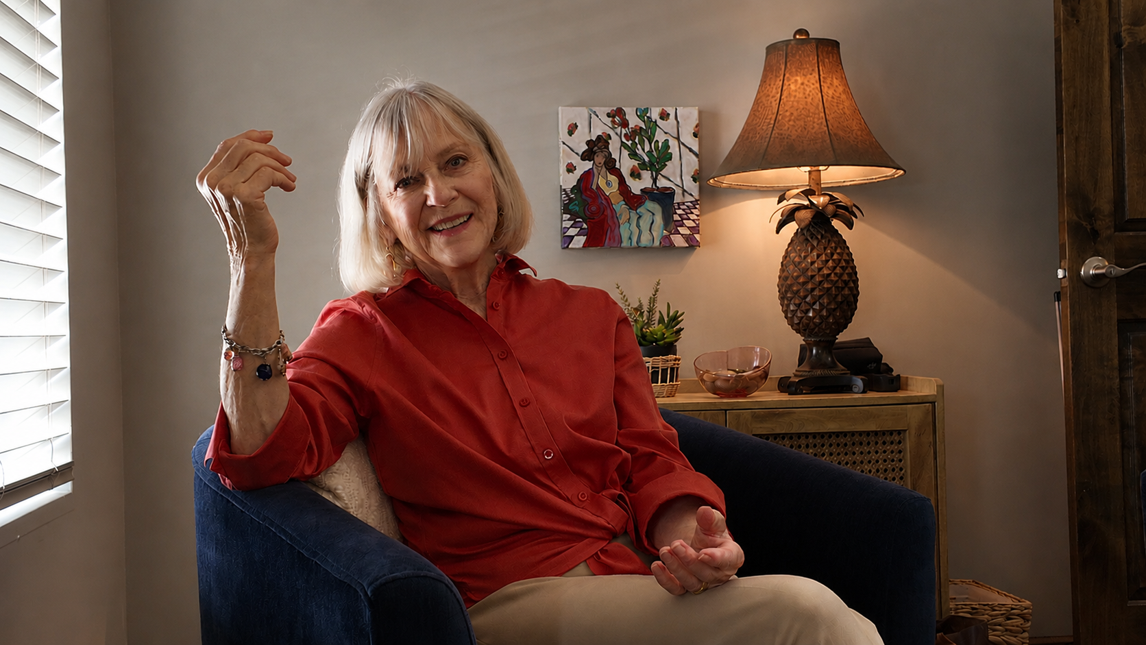 An older woman with blonde hair smiling and sitting in a blue armchair in a cozy living room. She is wearing a red blouse and bracelets. There is a window with blinds on the left, a painting on the wall behind her, and a lamp shaped like a pineapple on a wooden side table.