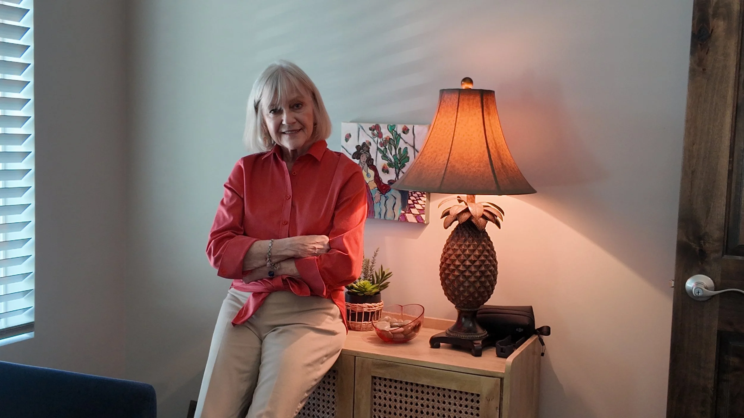 An older woman with blonde hair, wearing a red blouse and beige pants, standing with arms crossed in a room with modern decor. Behind her is a lamp with a pineapple-shaped base, a small painting with floral theme, and a plant on a wooden side table.