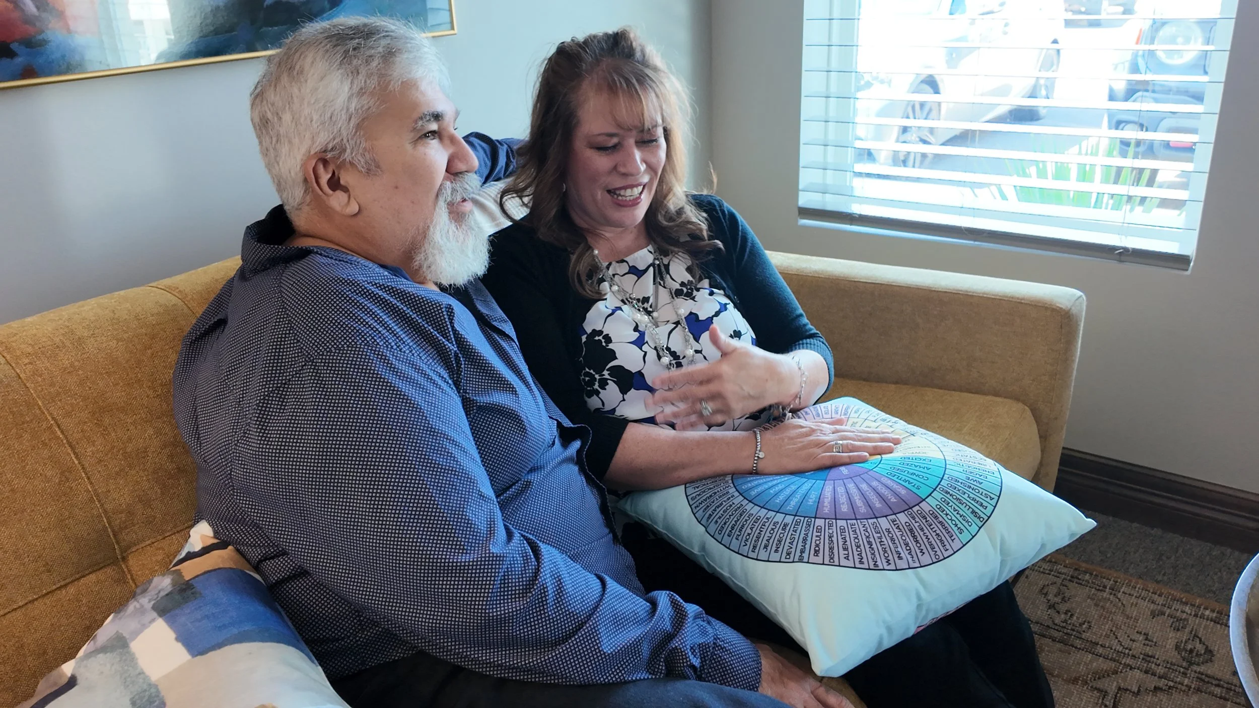 A man with gray hair and a beard sitting close to a woman with long, wavy hair. They are sharing an emotional moment, with the woman appearing to cry or be moved. They are sitting on a mustard-colored couch near a window with blinds. The woman is holding a folder or chart with various color-coded sections and text, resting on a pillow.