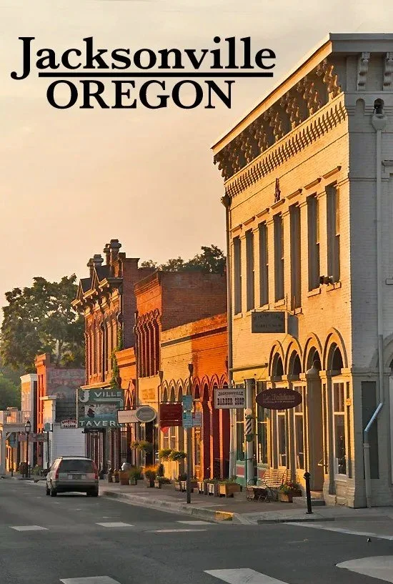 A street scene of historic buildings in Jacksonville, Oregon, during sunset with storefront signs and parked cars.
