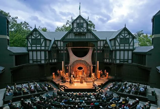 Outdoor theater stage with actors performing in front of a large round set piece, audience seated in front, surrounded by Tudor-style buildings with trees and a cloudy sky in the background.