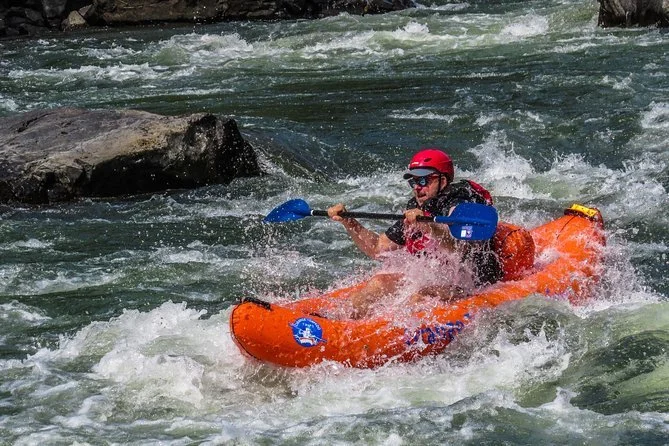Person kayaking on a river with rocks, wearing a red helmet and sunglasses, in an orange kayak.