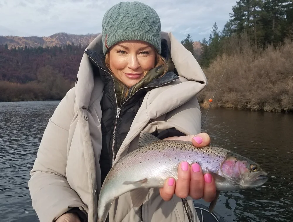 A woman in winter clothing holding a fish she caught, standing near a river with a hilly, wooded landscape in the background.