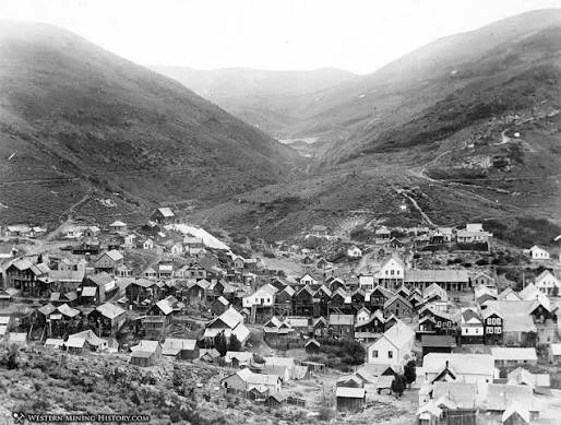 Black and white photo of a small mountain town with numerous wooden houses and buildings, surrounded by mountains in the background.