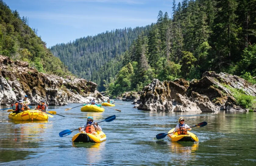 A group of people kayaking on a river surrounded by rocky banks and dense green forest under a clear blue sky.