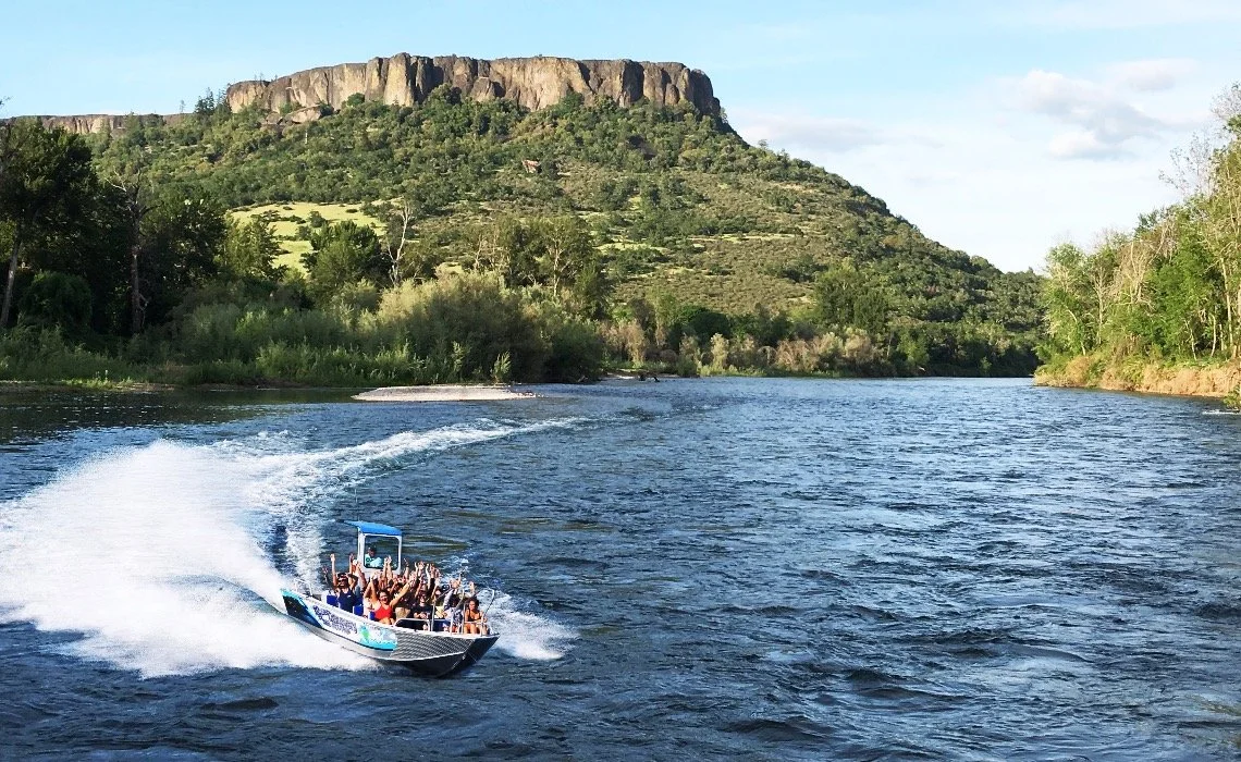 A boat with several passengers riding on a river surrounded by lush green trees with a rocky hill in the background under a partly cloudy sky.
