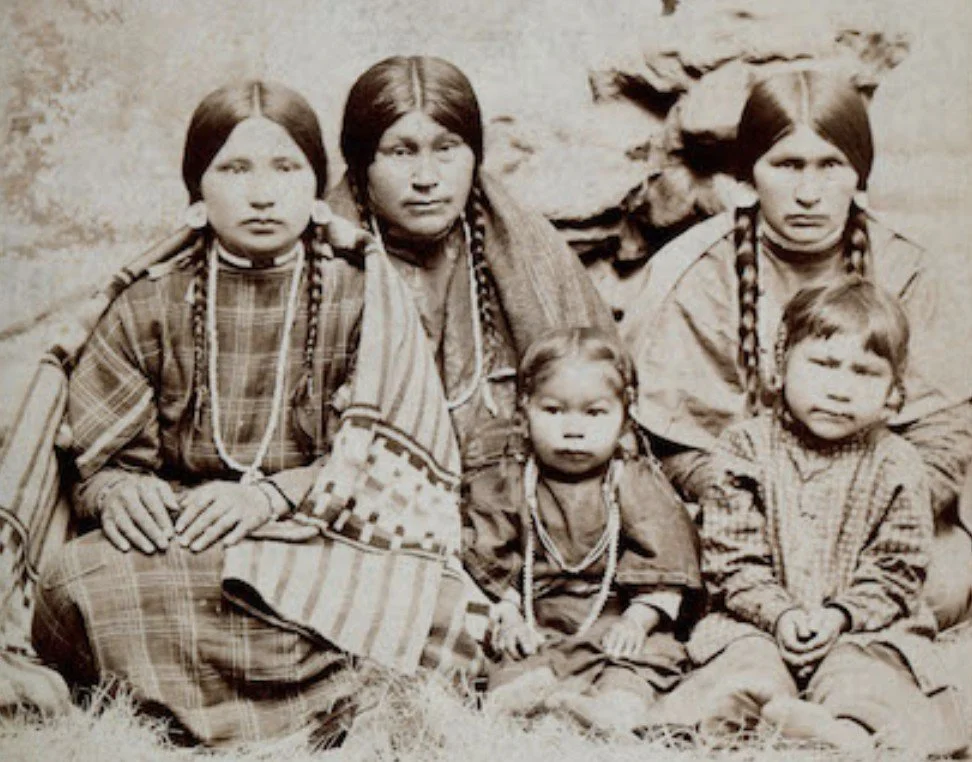 Five indigenous children sitting outdoors, dressed in traditional clothing, with a rocky background.