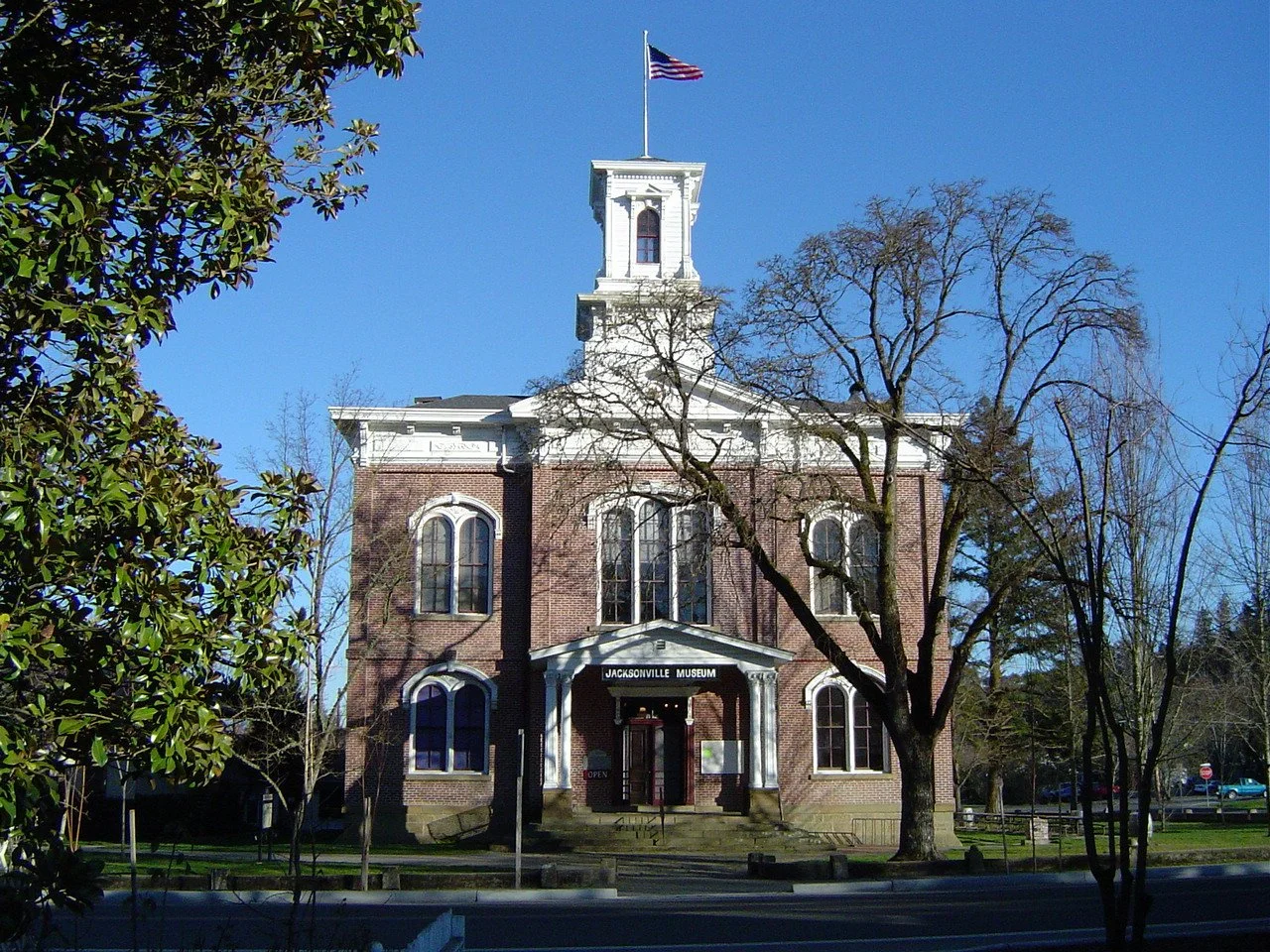 A historic brick building with a white steeple and a flag on top, labeled as Jacksonville Museum, surrounded by trees and a clear blue sky.