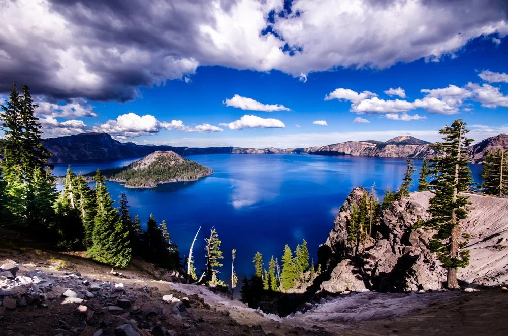 A volcanic crater lake with deep blue water surrounded by forested slopes and mountains under a partly cloudy sky.