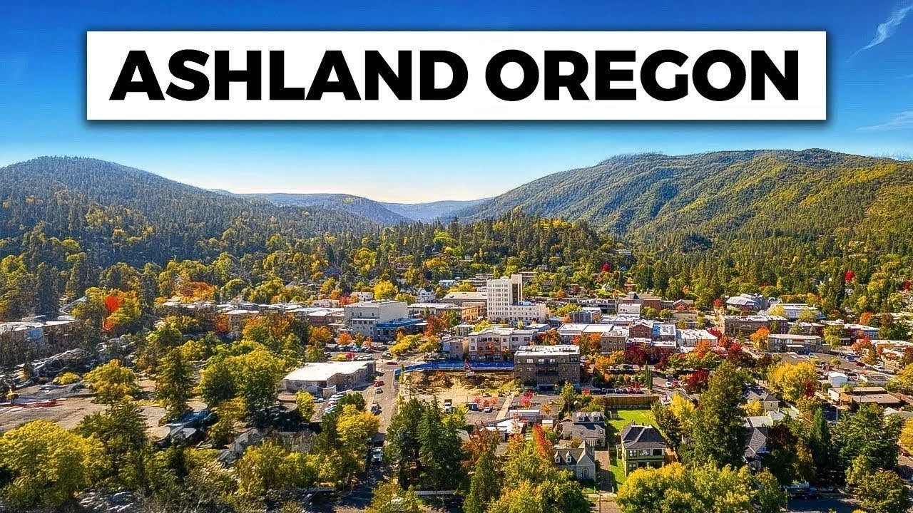 Aerial view of Ashland, Oregon, with small town buildings surrounded by lush green mountains and trees under a bright blue sky.