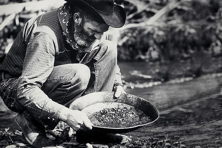 A man wearing a cowboy hat, bandana, denim jacket, and jeans is sitting by a creek, holding a pan and sifting through gravel and water for gold.