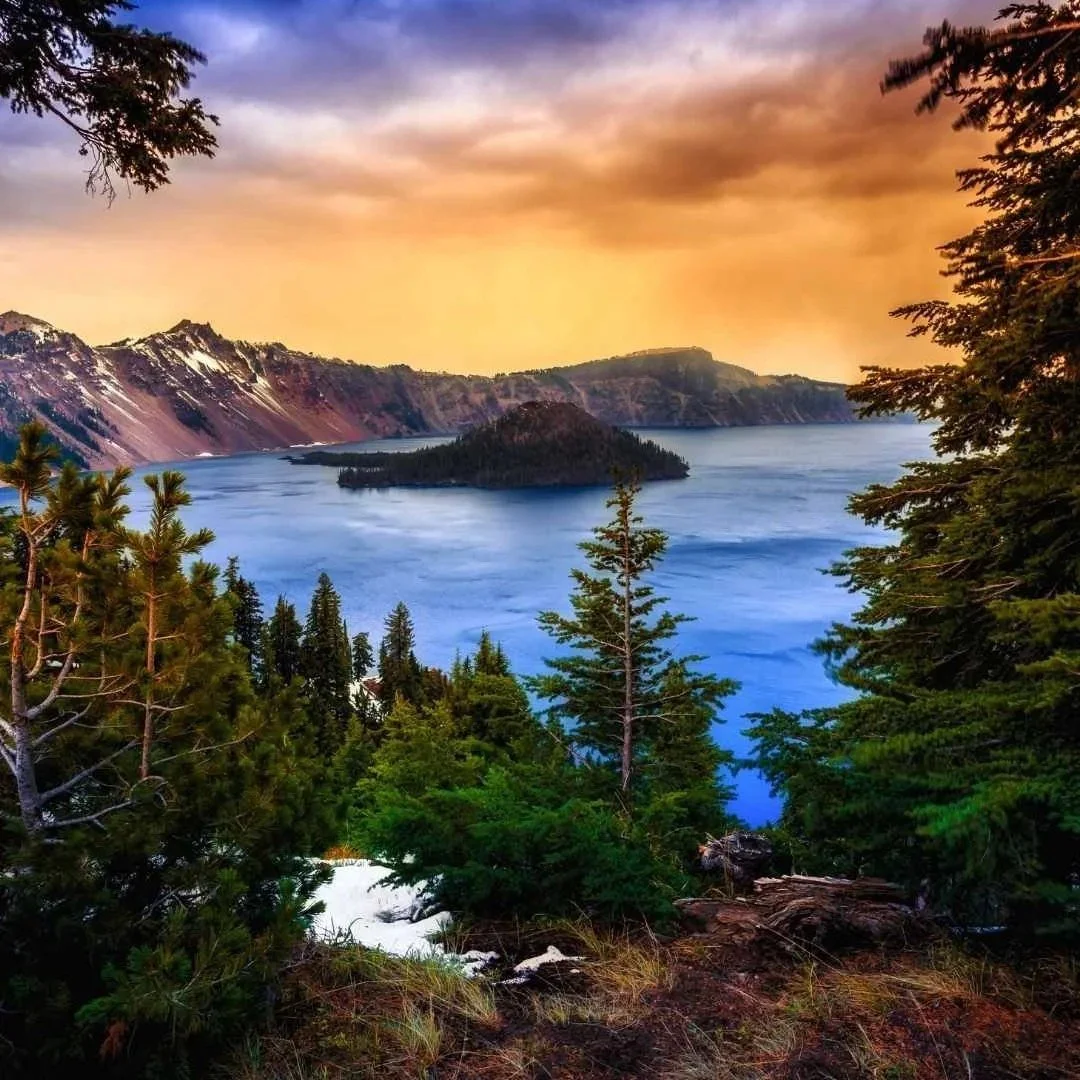 A scenic view of a lake with an island in the middle, surrounded by forested hills and mountains, with a colorful sky at sunset.