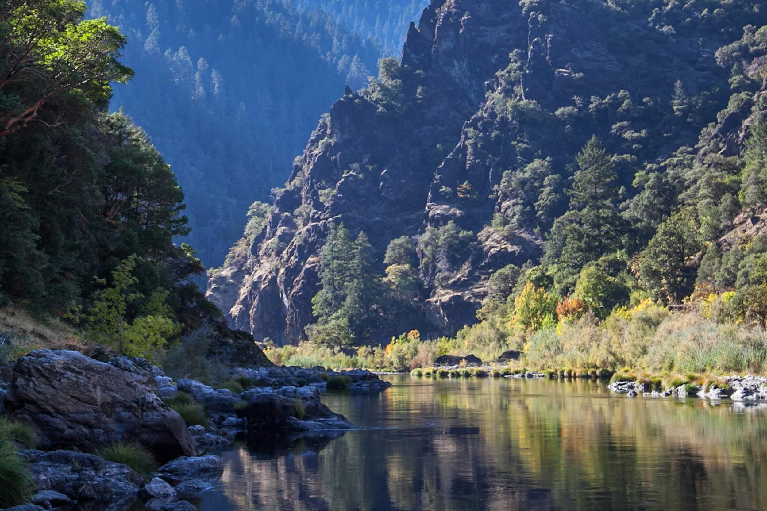 A scenic landscape featuring a calm river flowing through a forested area with lush green trees, surrounded by steep mountain cliffs under a clear blue sky.