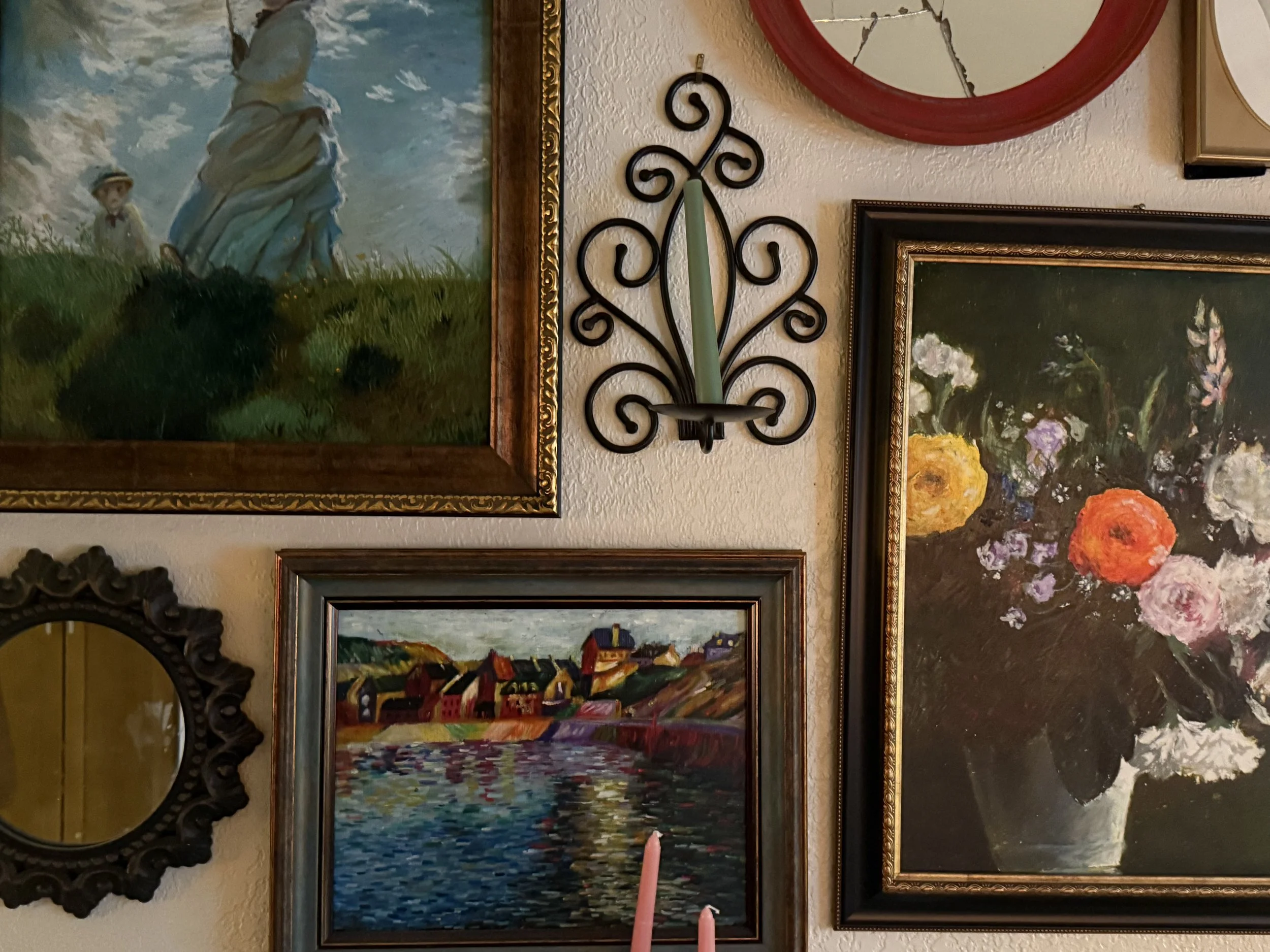 A wall decorated with framed paintings including a landscape, a cityscape, floral arrangements, and a woman, with a decorative wall clock and a mirror.