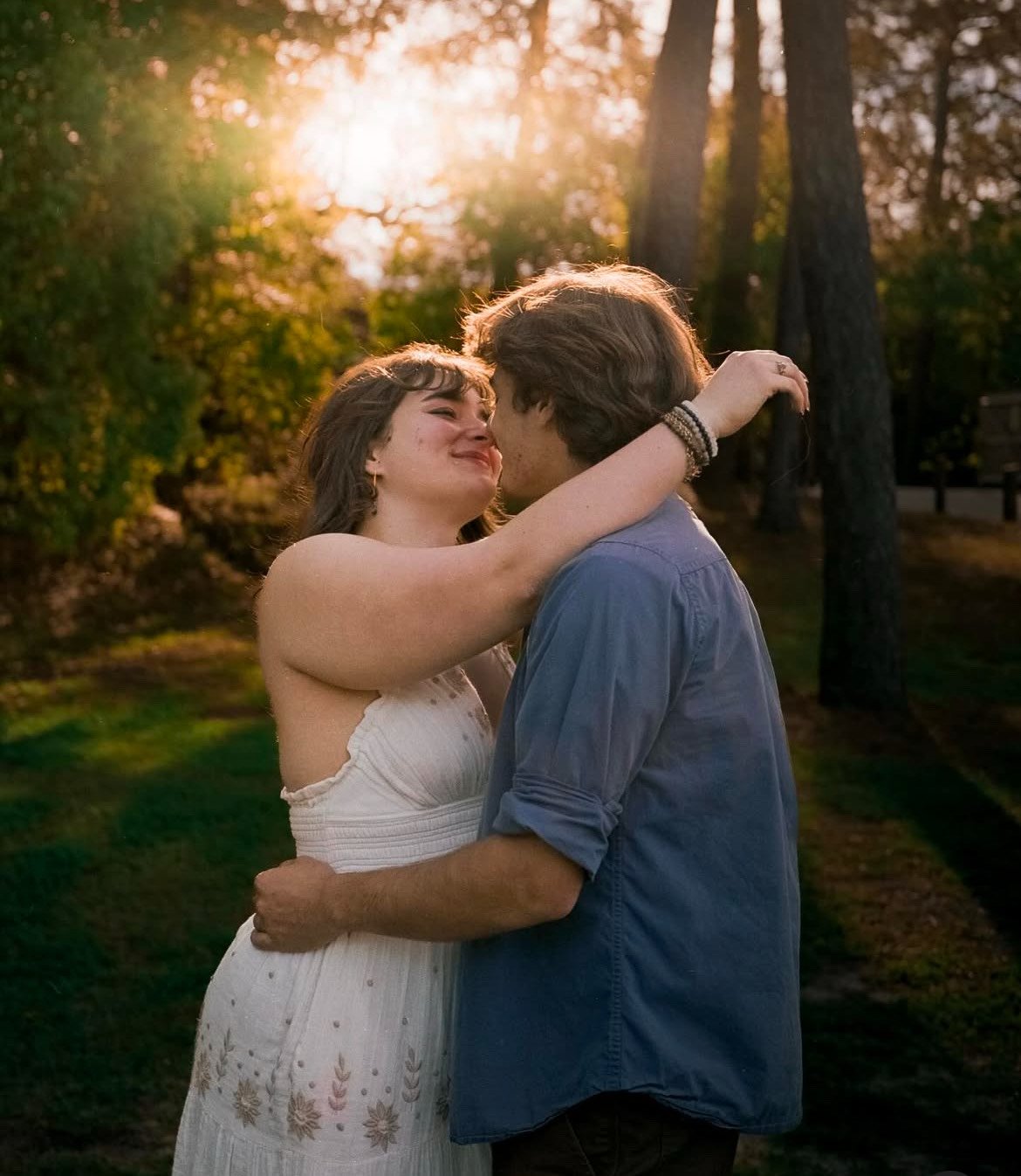 A couple embracing and touching noses in a park during sunset, with trees and sunlight in the background.