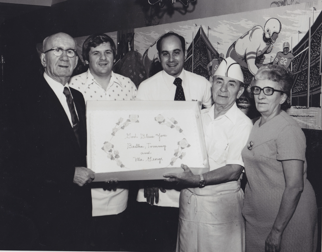 Five people standing together, holding a framed gift with a message, in front of a backdrop featuring football players and mountains, in black-and-white.