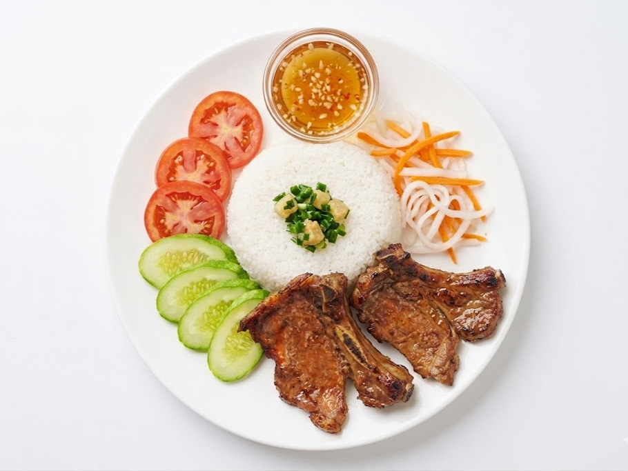 Plate with grilled pork chops, sliced cucumber and tomato, rice topped with chopped garlic and green onion, side of pickled vegetables, and a small bowl of dipping sauce.