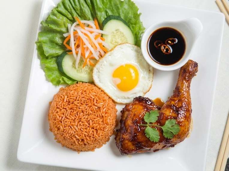 Plate of Asian-inspired meal with fried rice, grilled chicken drumstick glazed with sauce and garnished with cilantro, fried egg sunny side up, cucumber and carrot salad, and a small dish of soy sauce with sliced chilies.