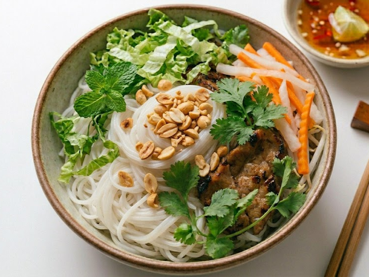 Bowl of rice noodles topped with chopped peanuts, fresh herbs, shredded carrots, sliced pickled vegetables, and grilled meat, with a side of dipping sauce.