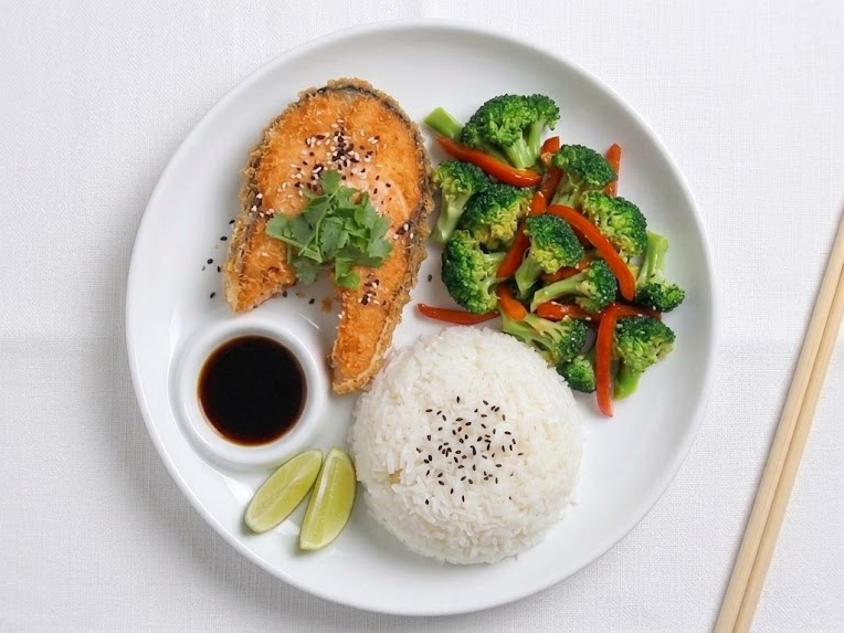 Fried fish fillet with garnished cilantro, served with white rice, steamed broccoli and red bell peppers, soy sauce, and lime wedges on a white plate.