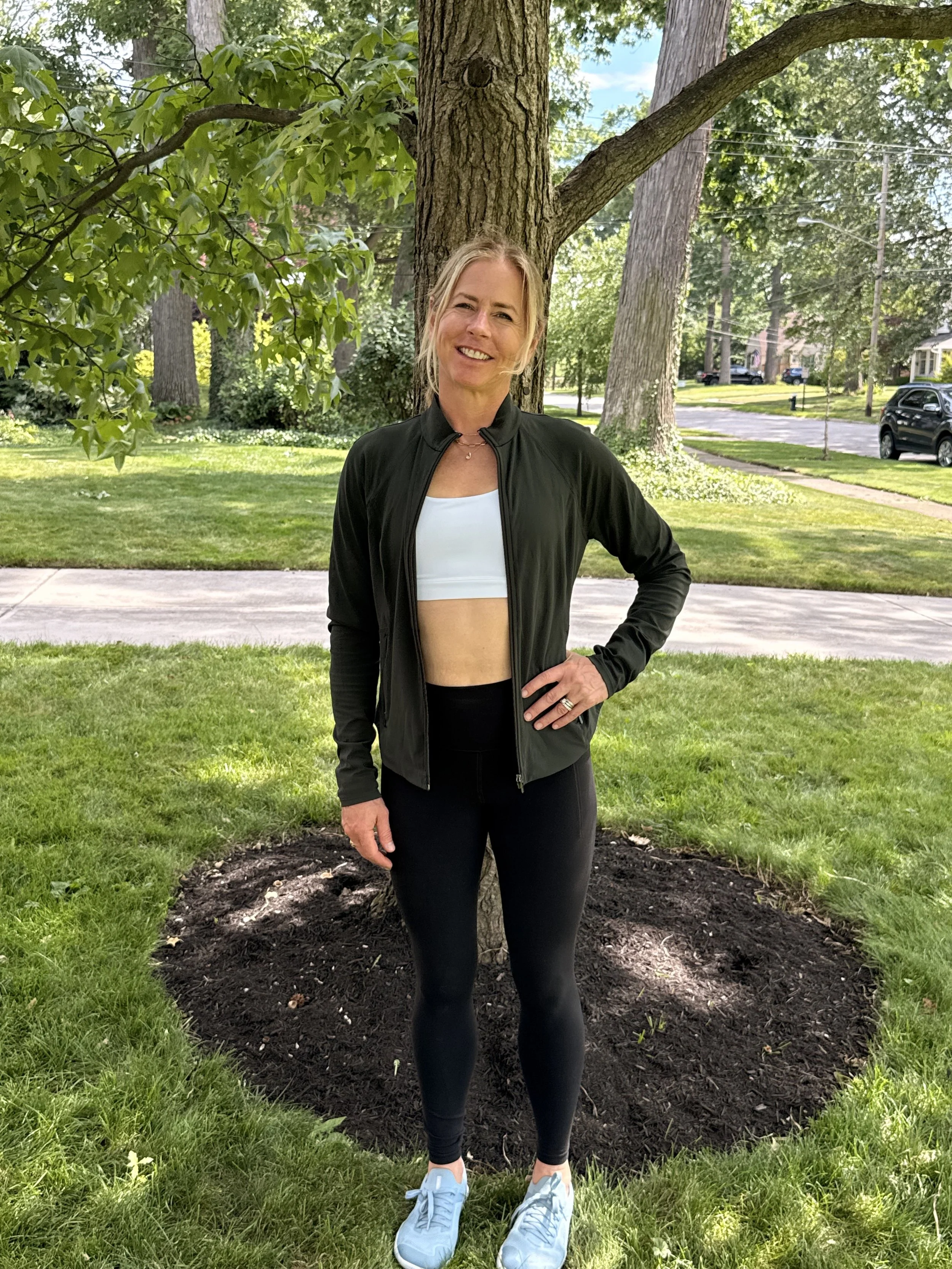 A woman with blonde hair tied back, wearing a black jacket, white sports bra, black leggings, and light blue sneakers, standing outdoors in a suburban neighborhood park with grass, trees, and houses in the background.