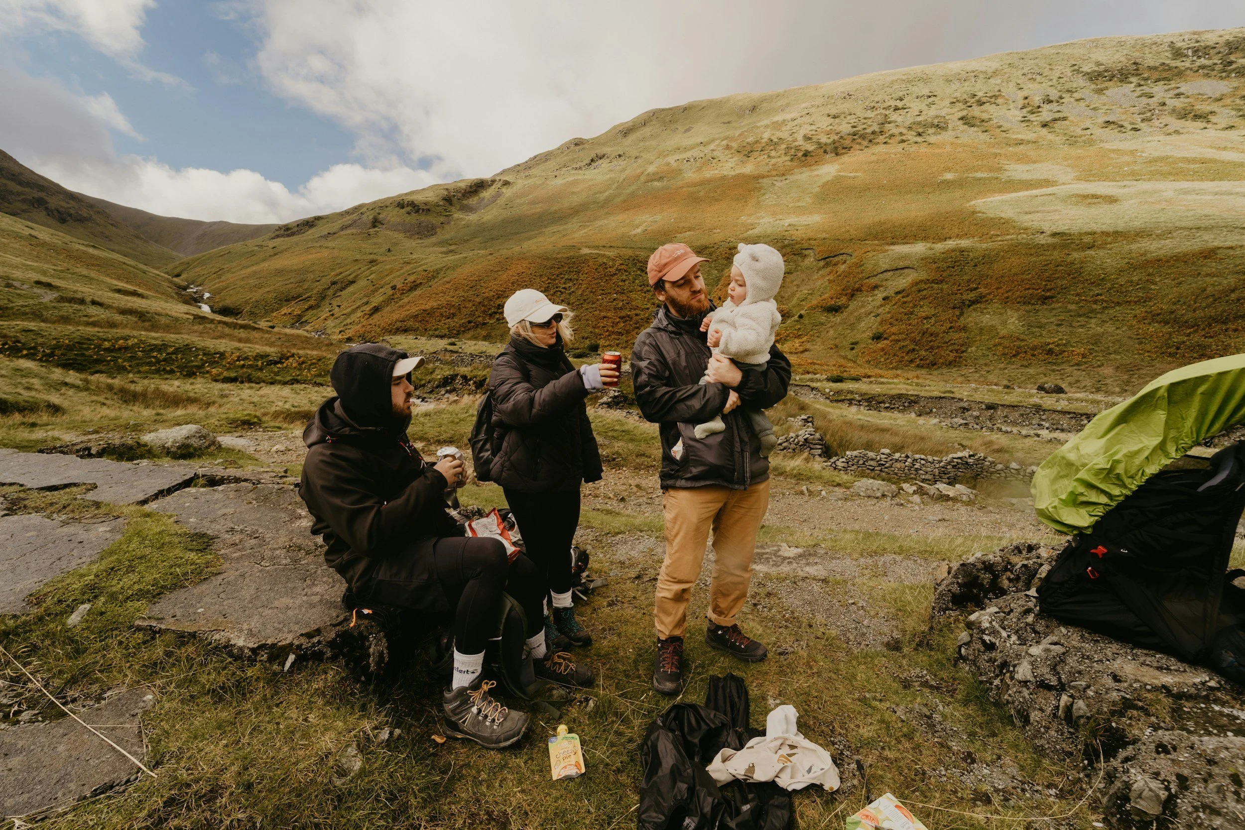 A family of four outdoors in a mountainous area with green hills and cloudy sky, preparing for camping. One person is sitting, the woman is standing holding a drink, the man is holding a young child, and they are all dressed warmly with hiking gear and jackets.