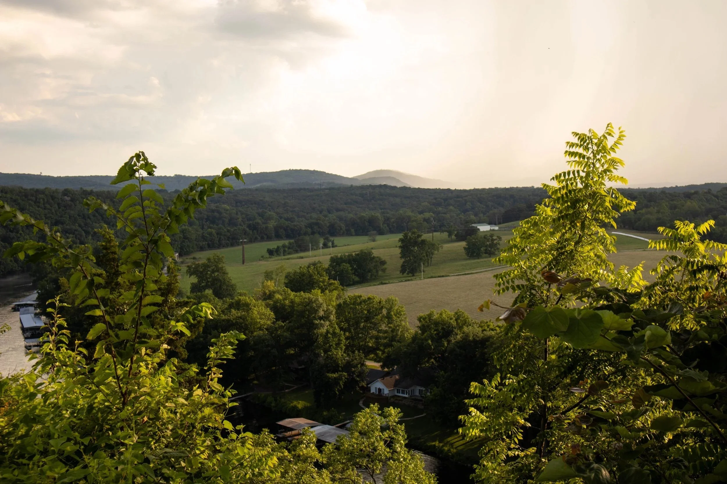 A scenic view of rolling green hills, trees, and a cloudy sky with some sunlight breaking through, overlooking a rural landscape with houses and a river.