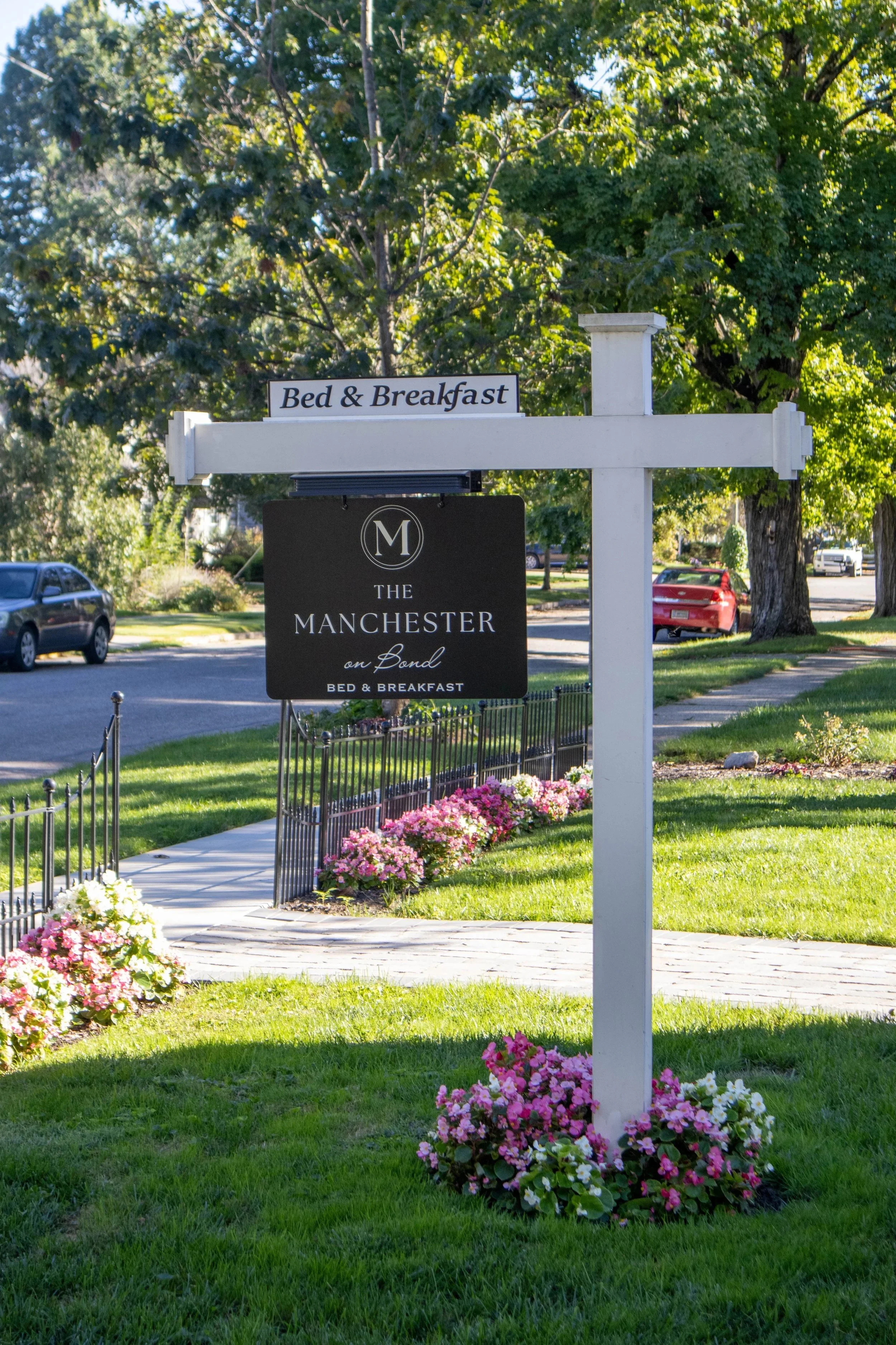 Sign for The Manchester Bed & Breakfast with pink and white flowers along a sidewalk, green grass, trees, parked cars in the background, and a clear sky.