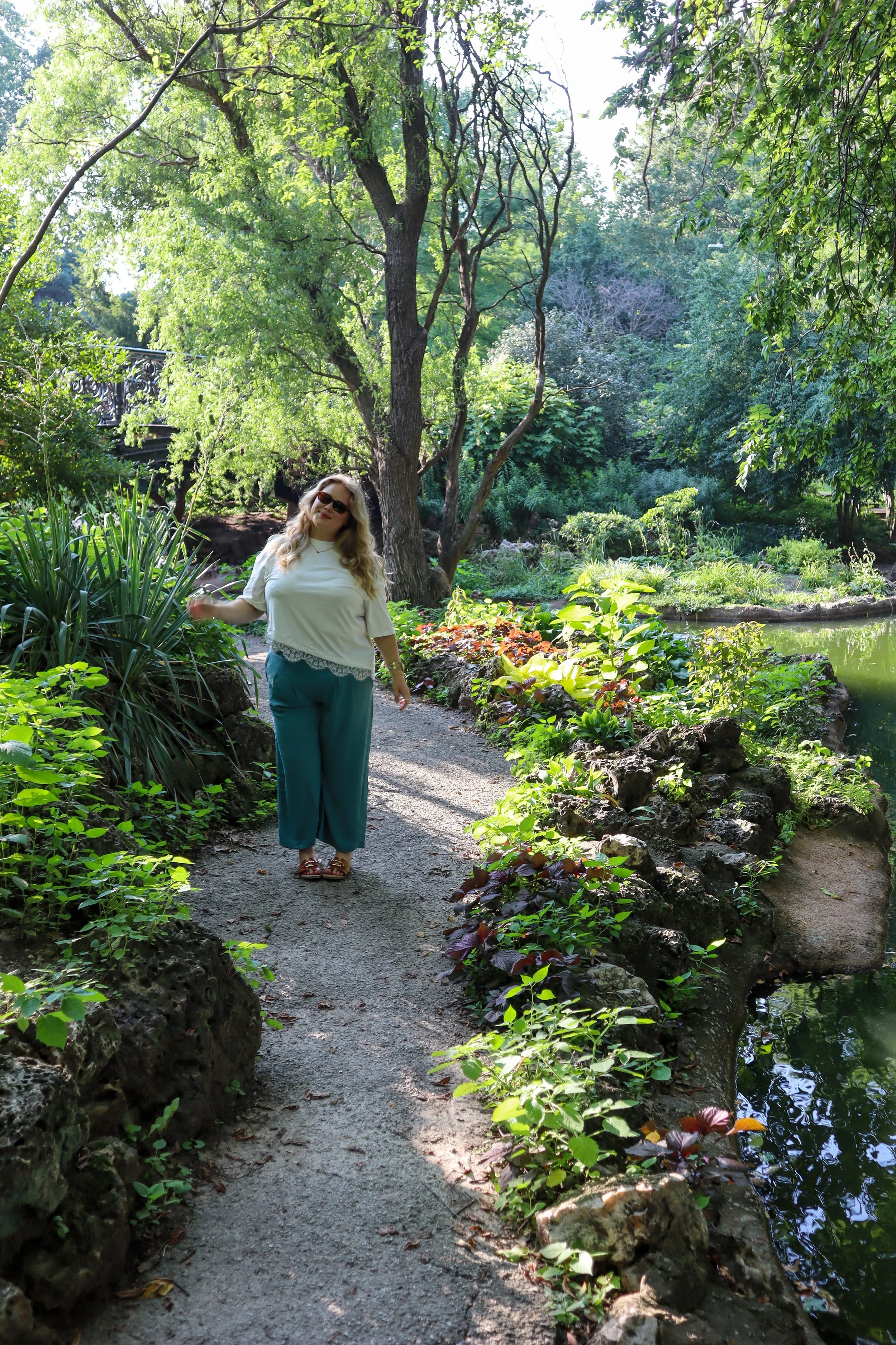 A woman with long blonde hair, wearing sunglasses, a white blouse, and teal pants, standing on a garden pathway surrounded by lush green trees and plants, with a small pond nearby.