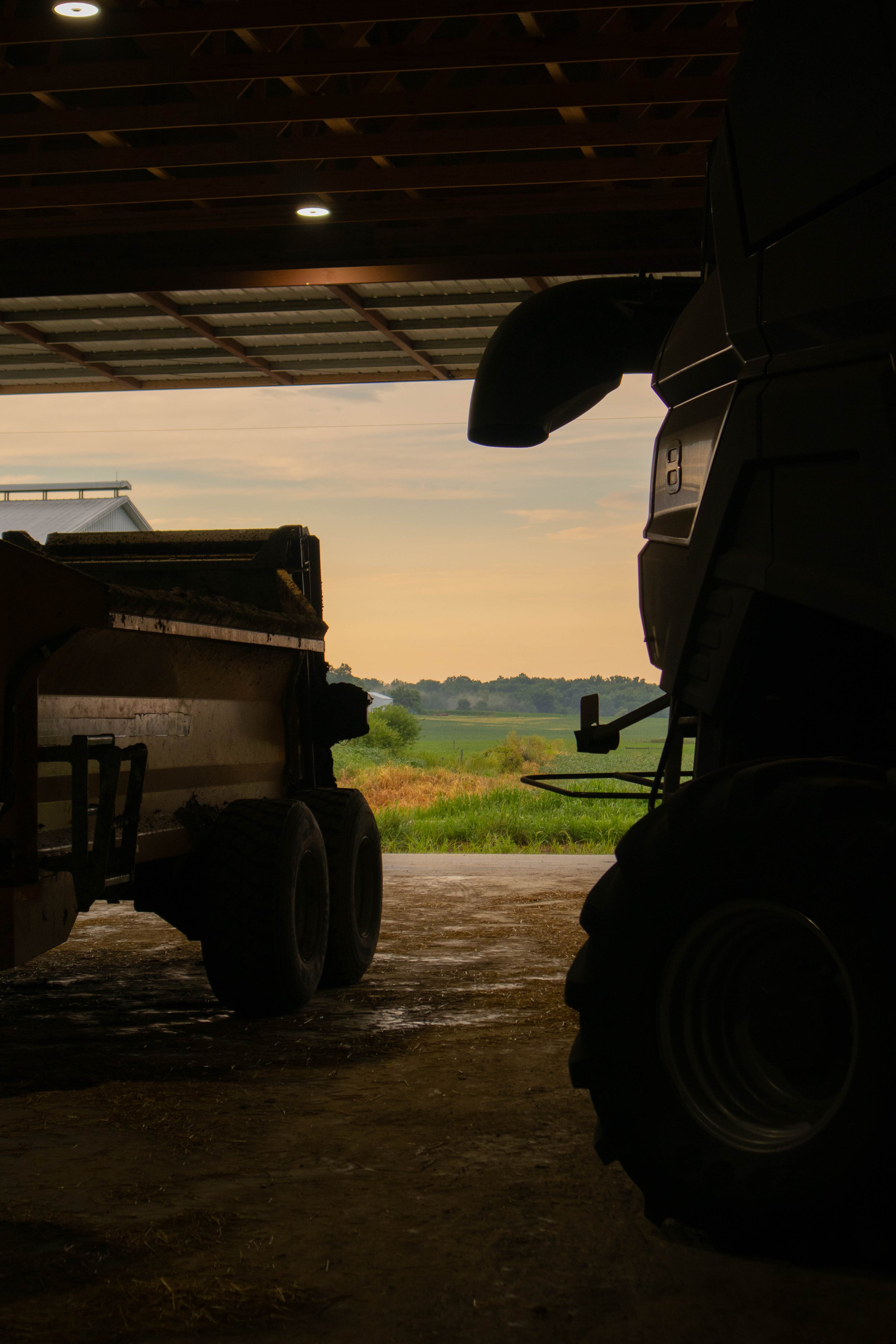 Silhouetted farm machinery inside a barn with open doors, showing a rural landscape and sunset sky outside.