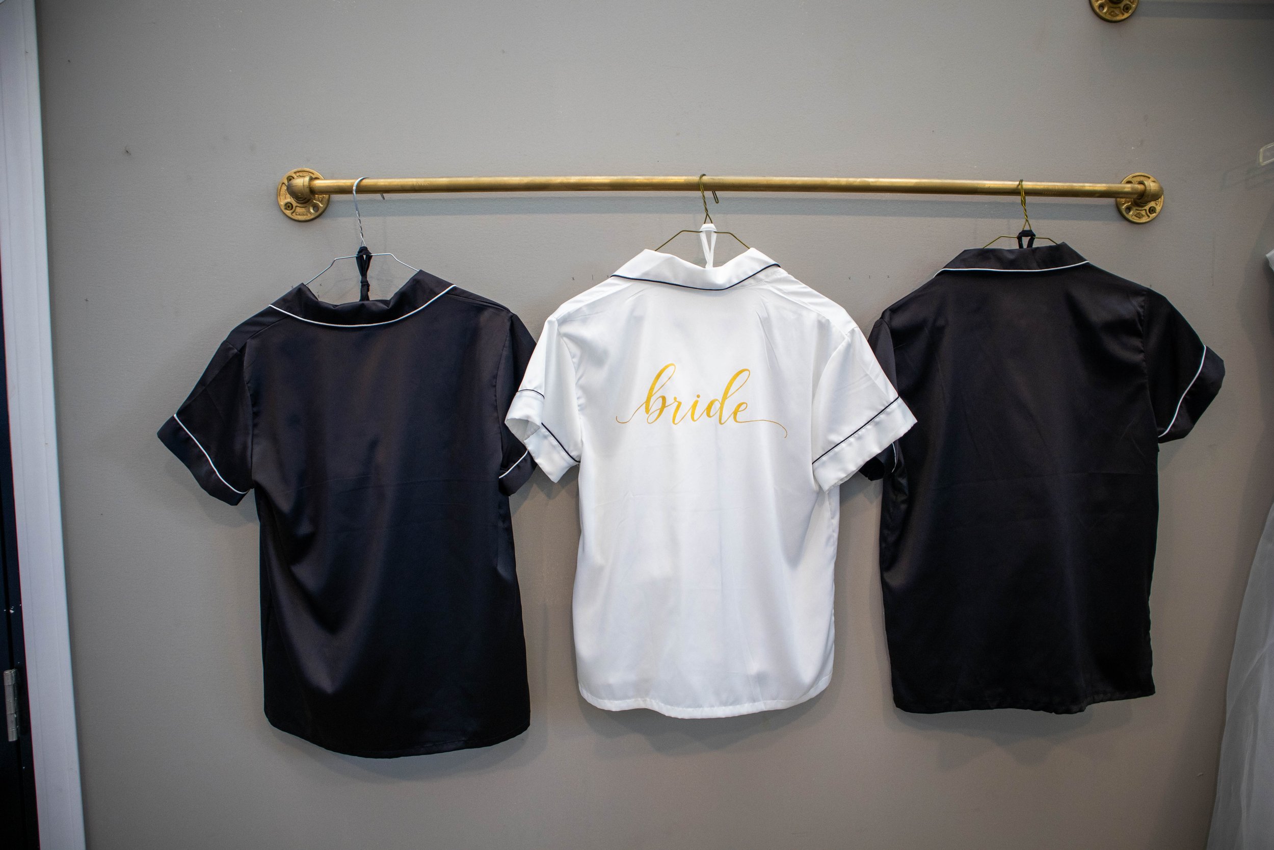 White robe with gold script that reads 'bride' hanging on a hanger, flanked by two black robes with white piping hanging on a gold-colored metal rod against a gray wall.