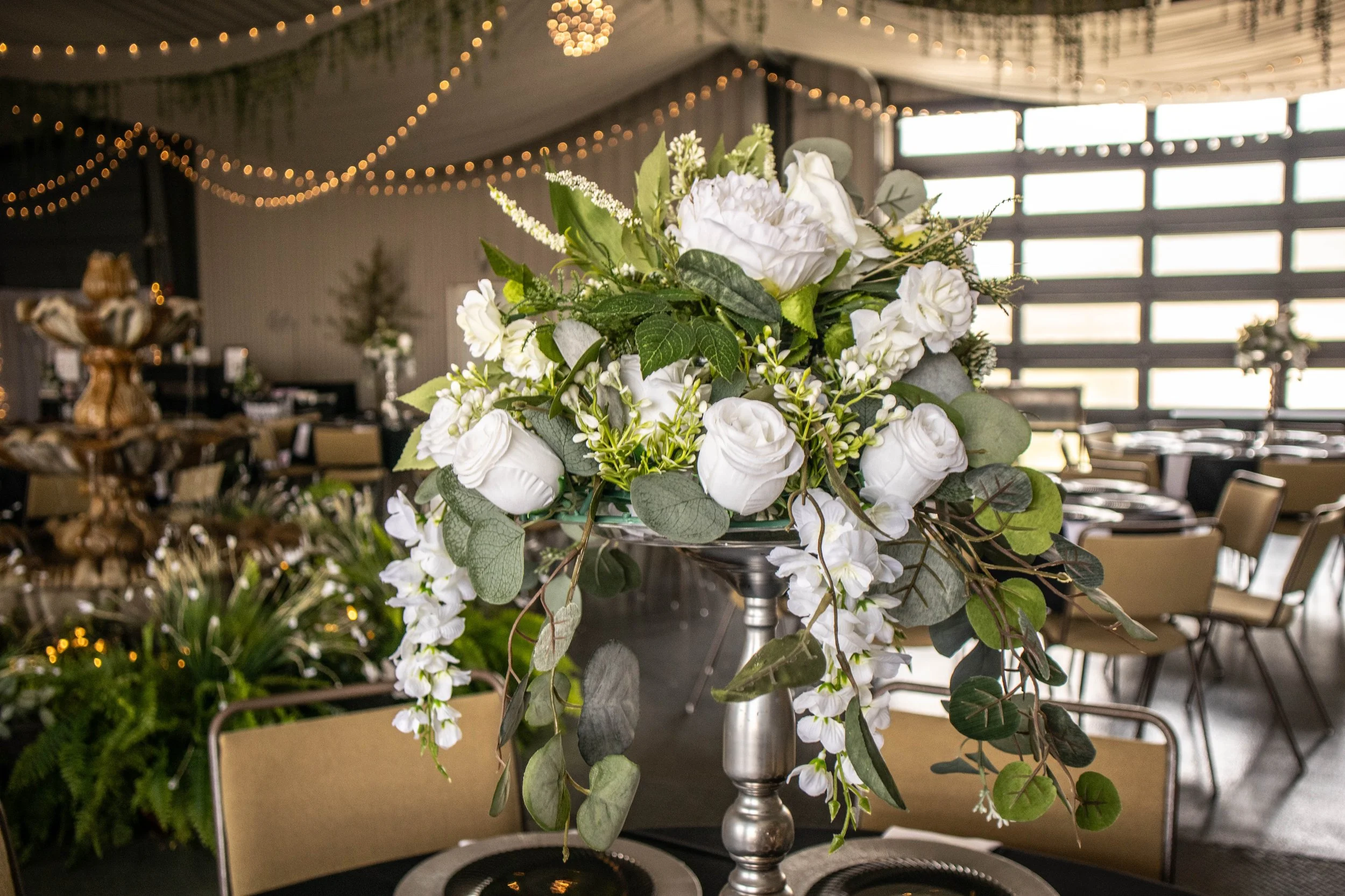 A tall floral centerpiece with white roses, green leaves, and other white flowers on a table at an event venue with string lights and large windows in the background