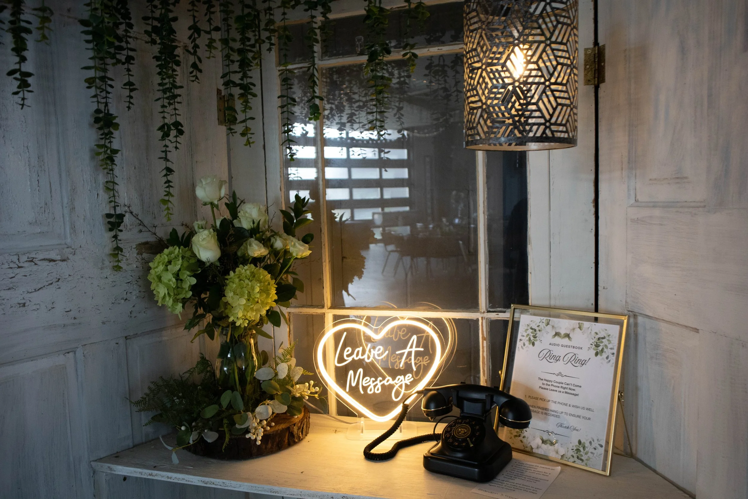 Decorative vintage-style setup with a bouquet of white roses and greenery, a neon heart sign with the words 'Leave a Message,' a black antique telephone, and a framed sign with instructions for leaving a message at a wedding or event. The background 